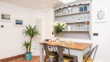 Un comedor con una mesa de madera, sillas metálicas, un florero con flores y una estantería con copas y frascos.