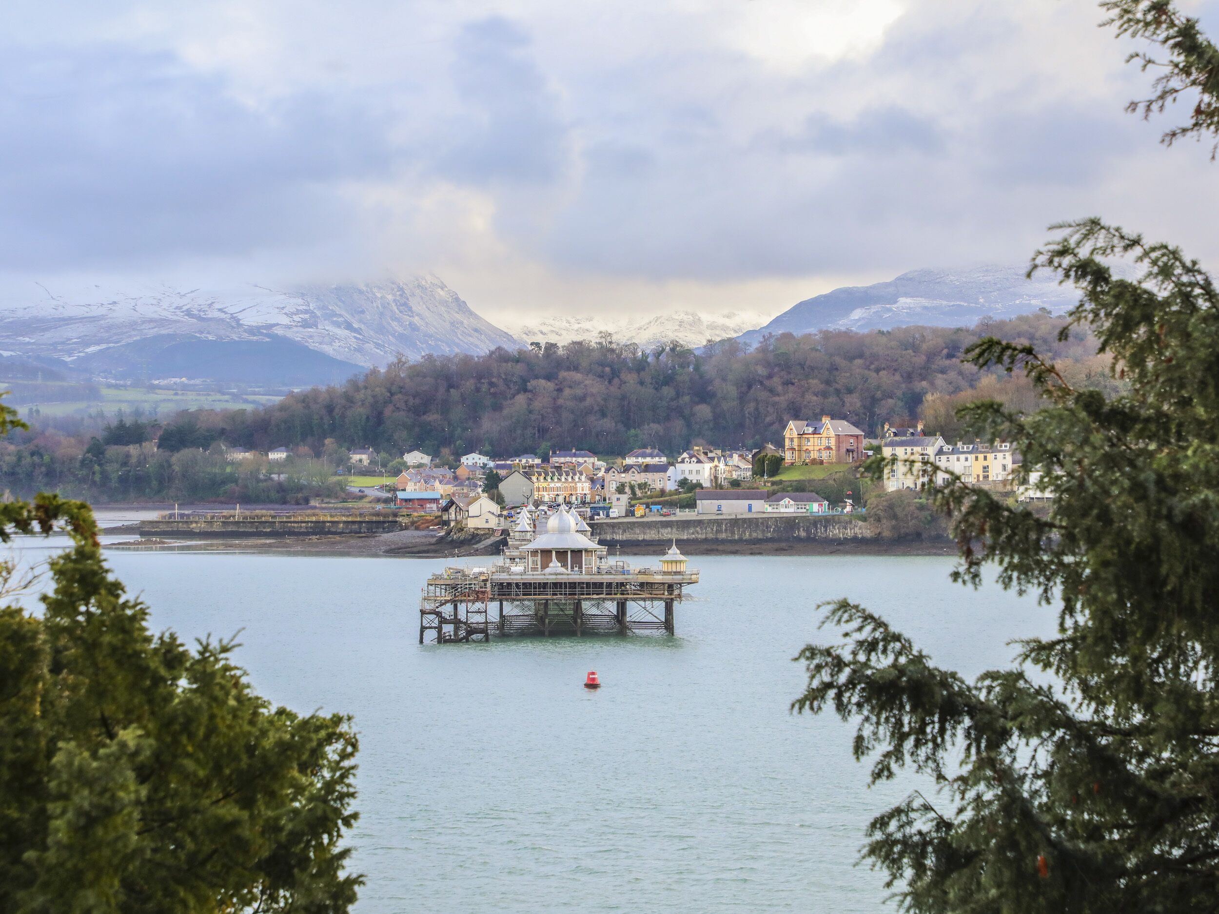 A lakeside town with a prominent pier structure in the foreground, surrounded by residential buildings and a backdrop of snow-capped mountains.