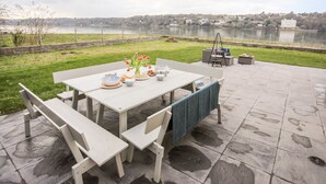 En udendørs terrasse med et spisebord, omgivet af stole og en bænk, med udsigt over en sø. I det fjerne ses et hegn og bygninger.