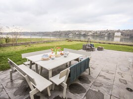 Un patio al aire libre con una mesa lista para comer, rodeado de sillas y un banco, con vistas a un lago con una cerca y edificios al fondo.