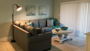 A living room with a grey sofa, a floor lamp, a round table with a green bowl, and two sailboat paintings on the wall. - Beautiful 2 bedroom 2 Bath directly across from Gulf (North Redington Beach)
