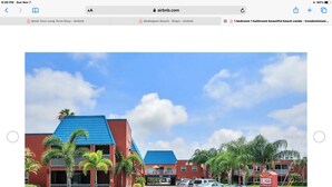 A modern building with a red facade and blue roof, surrounded by palm trees and parked cars. - Beautiful 2 bedroom 2 Bath directly across from Gulf (North Redington Beach)