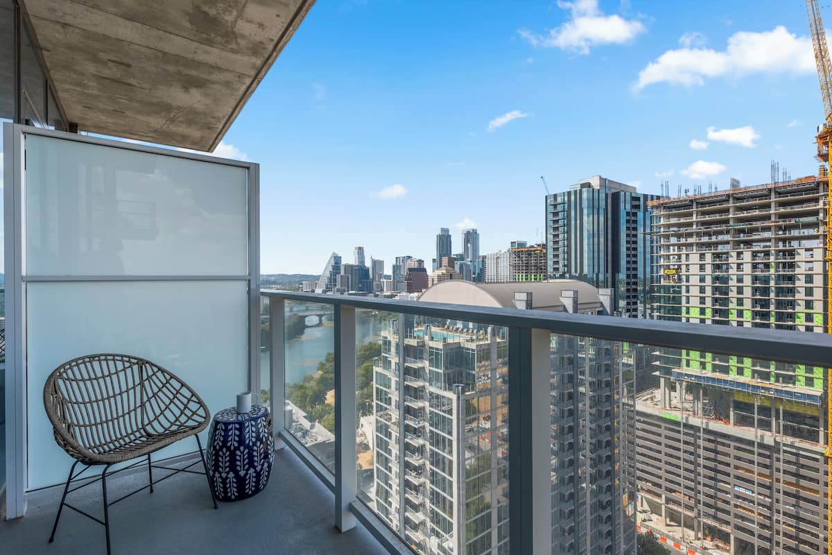 A balcony with a wicker chair and a patterned stool, overlooking a cityscape with modern buildings and construction sites.
