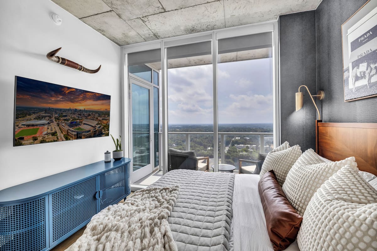 A bedroom with a large bed, a cityscape painting, a blue console, and a view of the city through large windows.