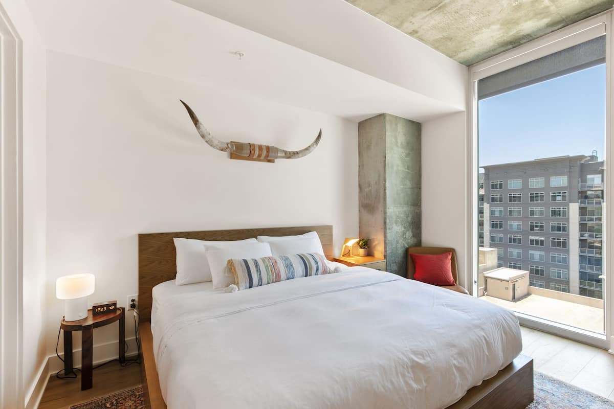 A modern hotel room with a large bed, a bedside table with a lamp, a decorative wall piece, and a view of the city through a large window.