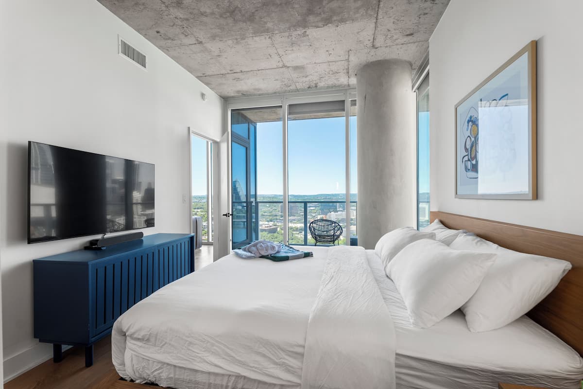 A modern bedroom with a large bed, a TV, a blue console, and a city view through large windows.
