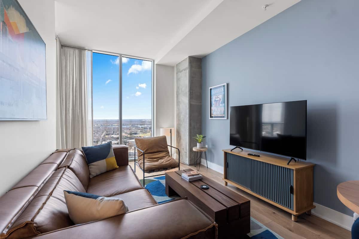A modern living room with a leather sofa, a wooden coffee table, a TV on a wooden stand, and a large window with a city view.