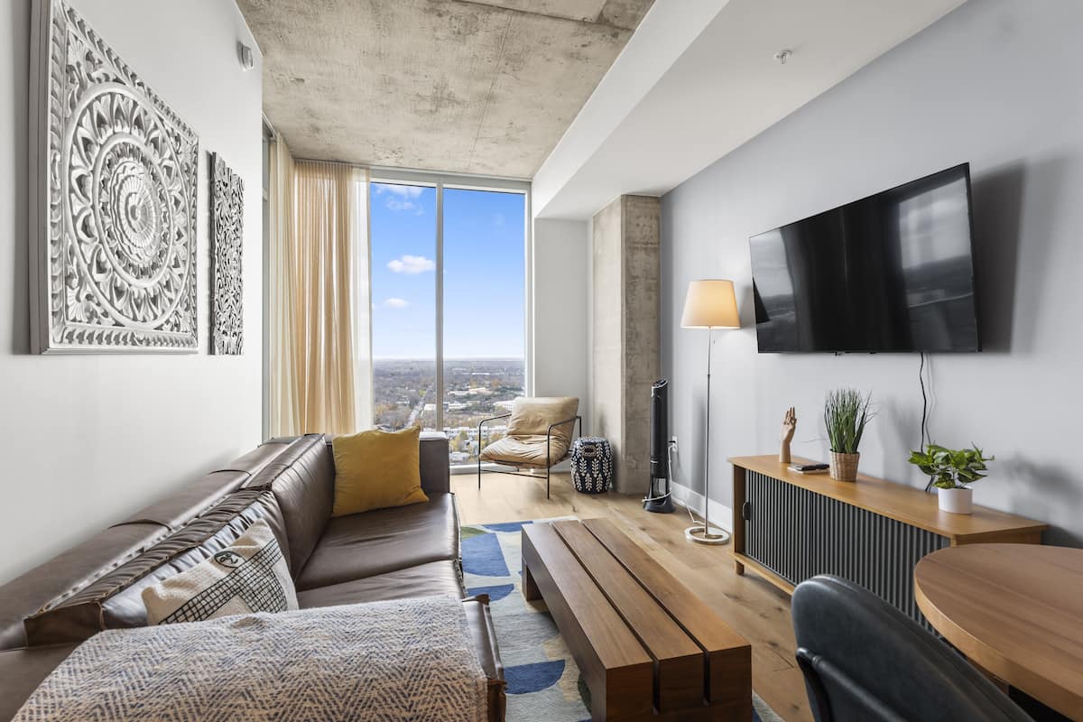 A modern living room with a sofa, a wooden coffee table, a TV mounted on the wall, and a view of the city through large windows.