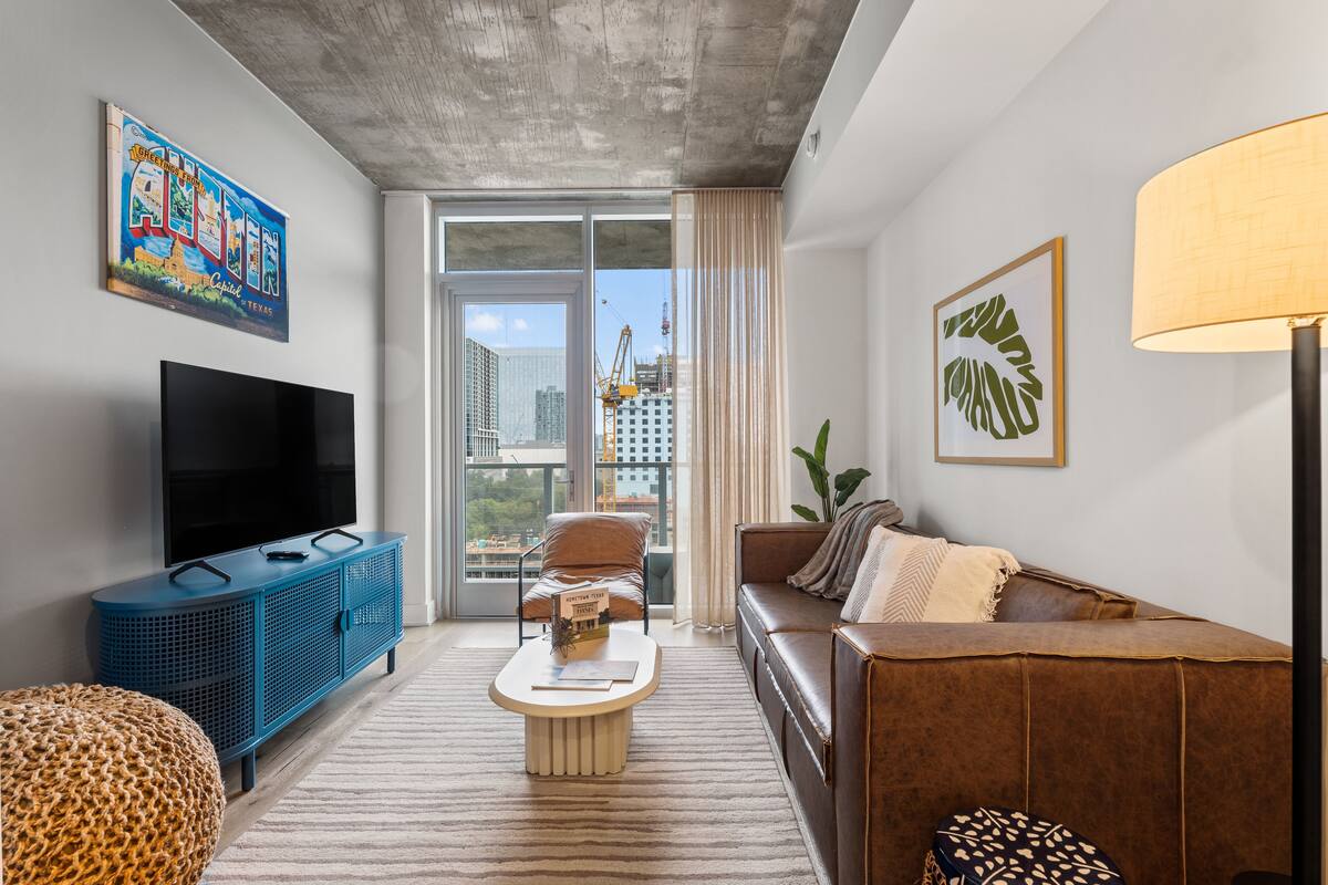 A modern living room with a large TV, a blue entertainment unit, a brown leather sofa, and a city view through the glass door.