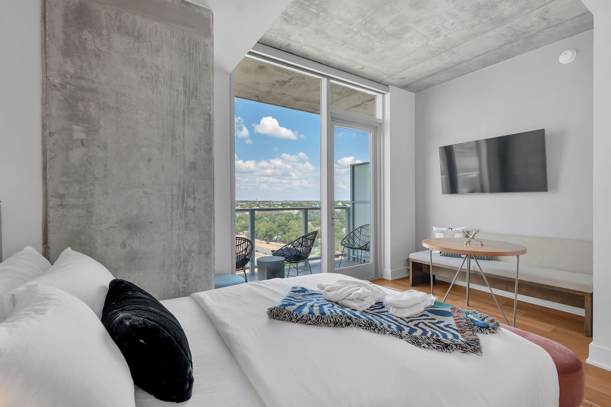 A modern hotel room with a large bed, a balcony with a view, a small round table, and a mounted TV.