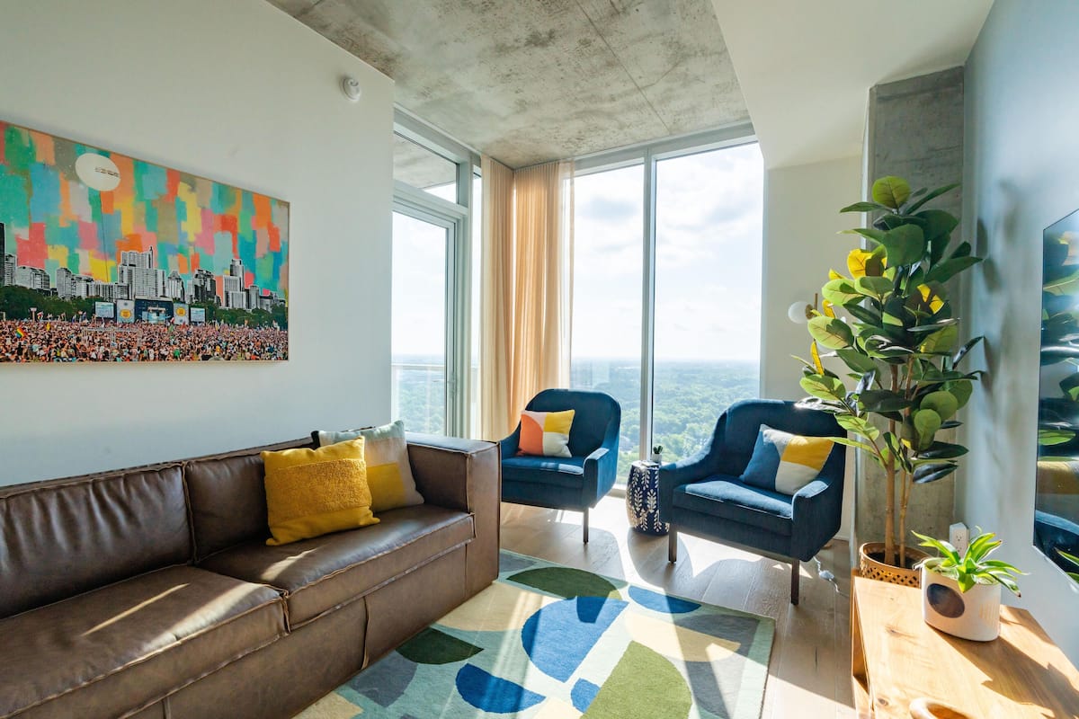 A modern living room with a brown leather sofa, two blue armchairs, a colorful abstract painting, and a large window offering a city view.