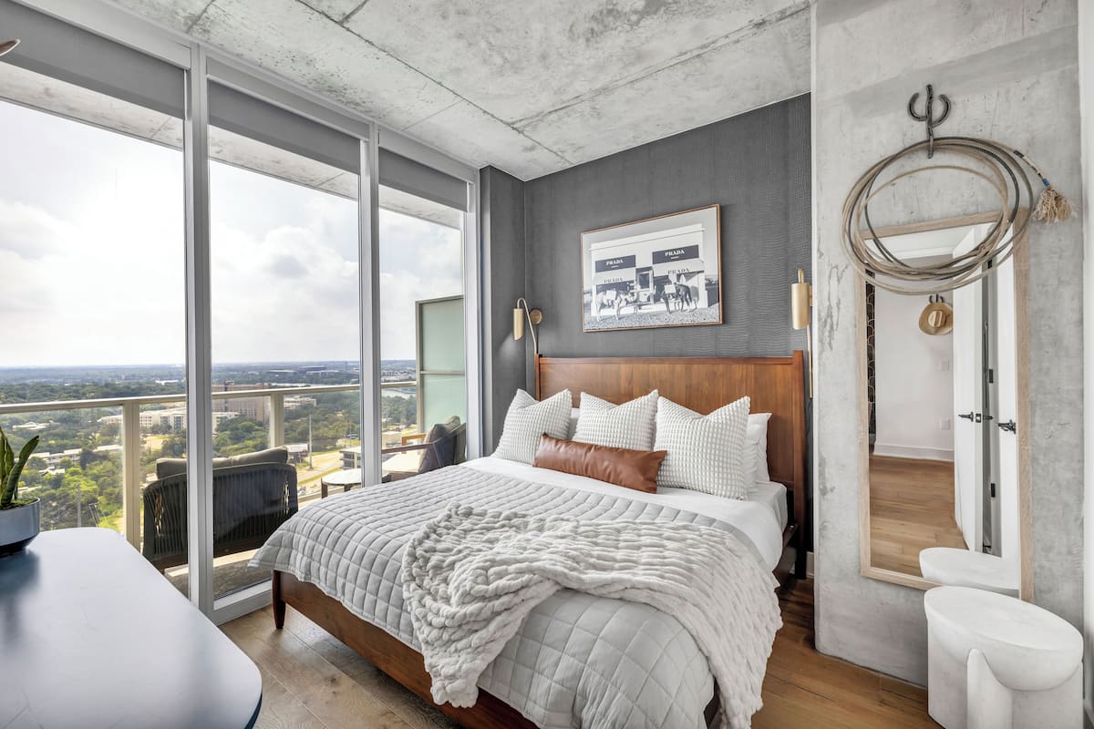 A modern bedroom with a large bed, a city view, a framed photo on the wall, and a unique mirror design.