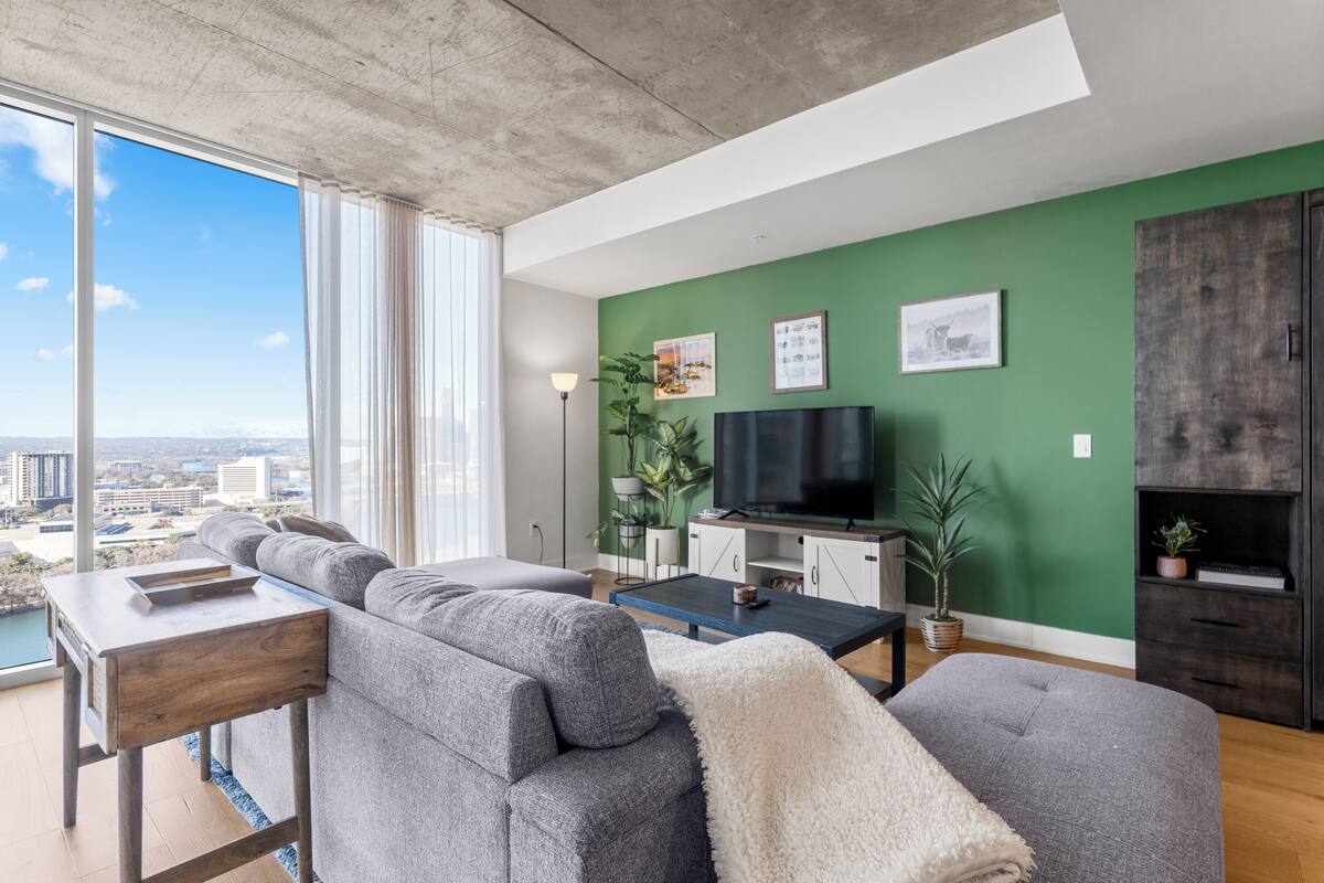 A modern living room with a large window, grey sofas, a flat-screen TV, and a green accent wall.