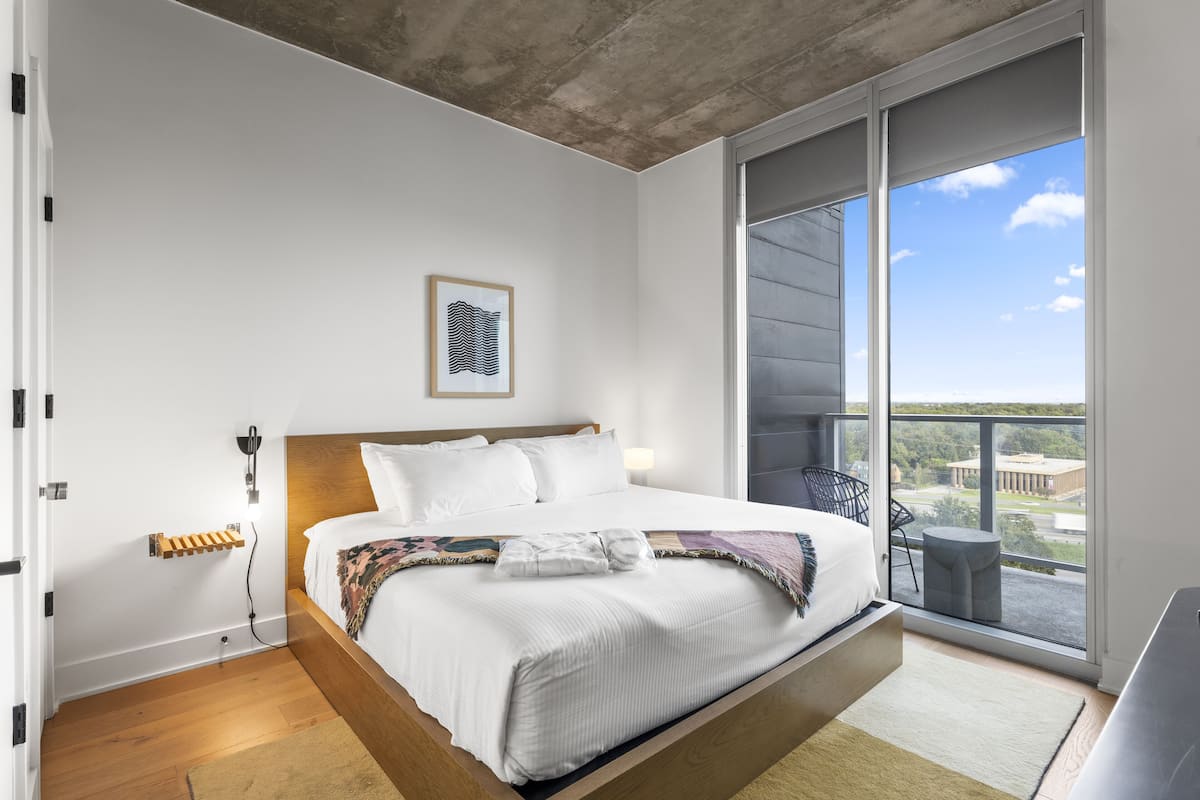 A modern bedroom with a bed, a bedside table, a wall-mounted light, and a large window with a view.