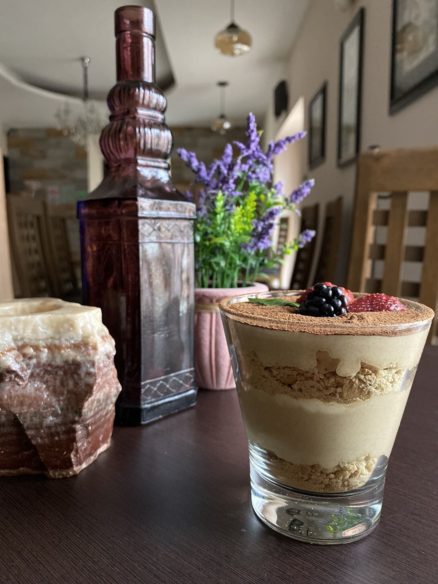 Un postre en un vaso sobre una mesa, con una botella decorativa y un jarrón de flores al fondo.