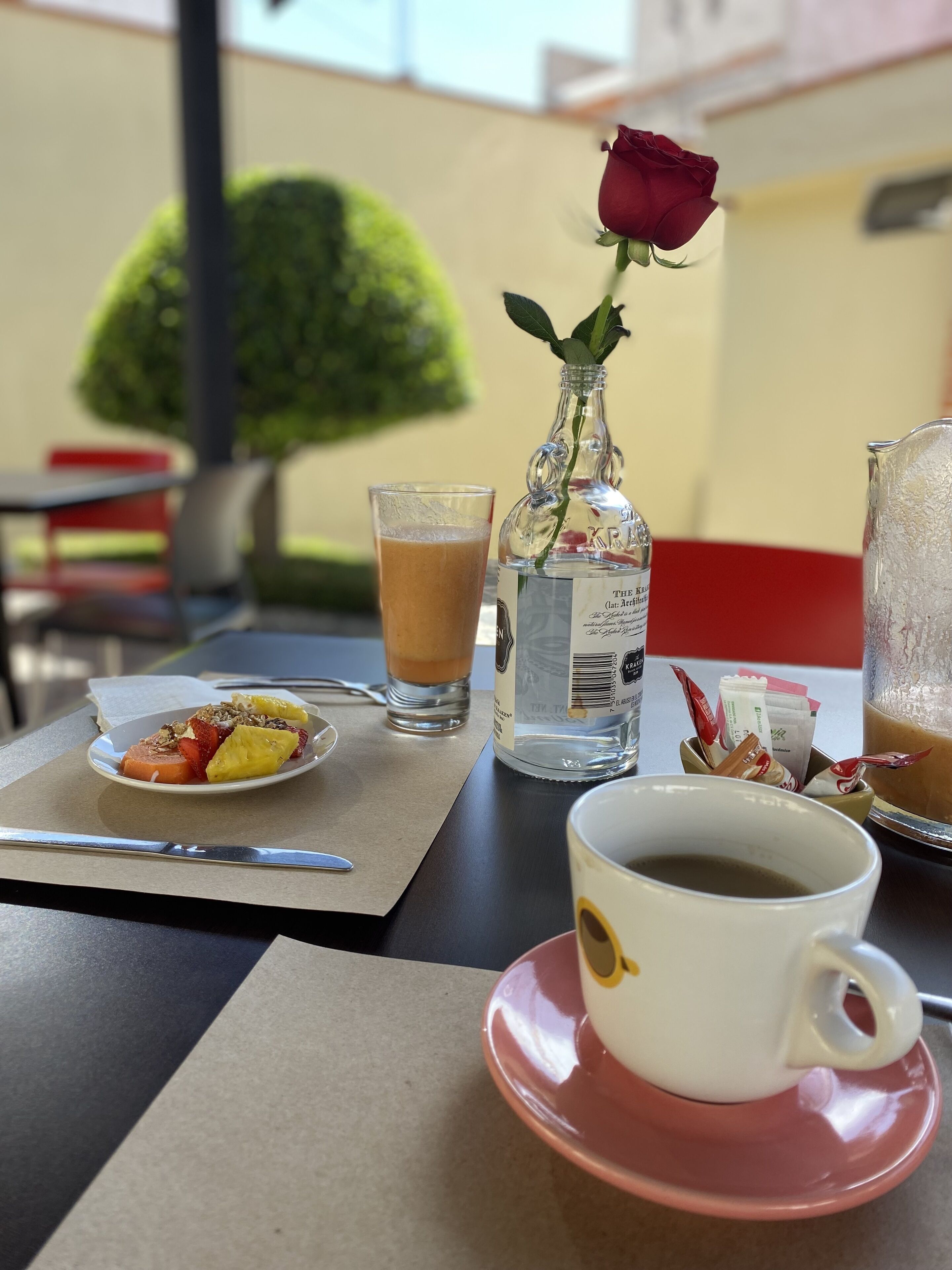 A table with a coffee cup, a glass of iced coffee, a bottle of water, a rose, and a plate of fruit and granola.