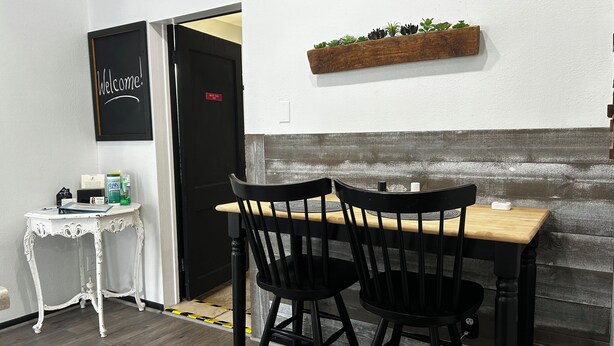 Ein kleiner Essbereich mit einem Holztisch und zwei Stühlen, ein weißer Nachttisch mit einigen Gegenständen, eine Tafel mit der Aufschrift 'Welcome!' und eine Holzablage mit Pflanzen.