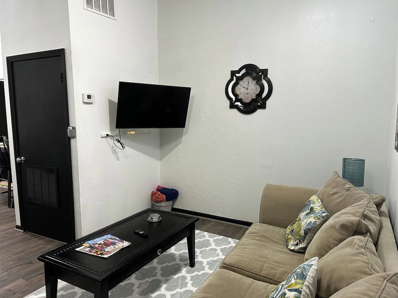 Un salon avec un téléviseur noir fixé au mur, une table basse noire sur laquelle sont posés des magazines, un canapé beige agrémenté de coussins et un tapis à motifs.