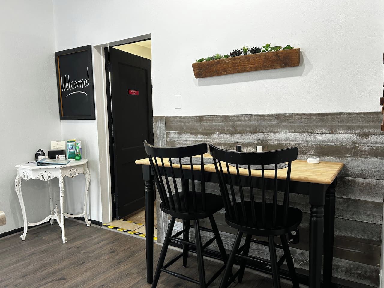 Una habitación con una mesa de madera y dos sillas negras, una mesa blanca con un objeto pequeño encima, y un letrero de bienvenida en la pared.