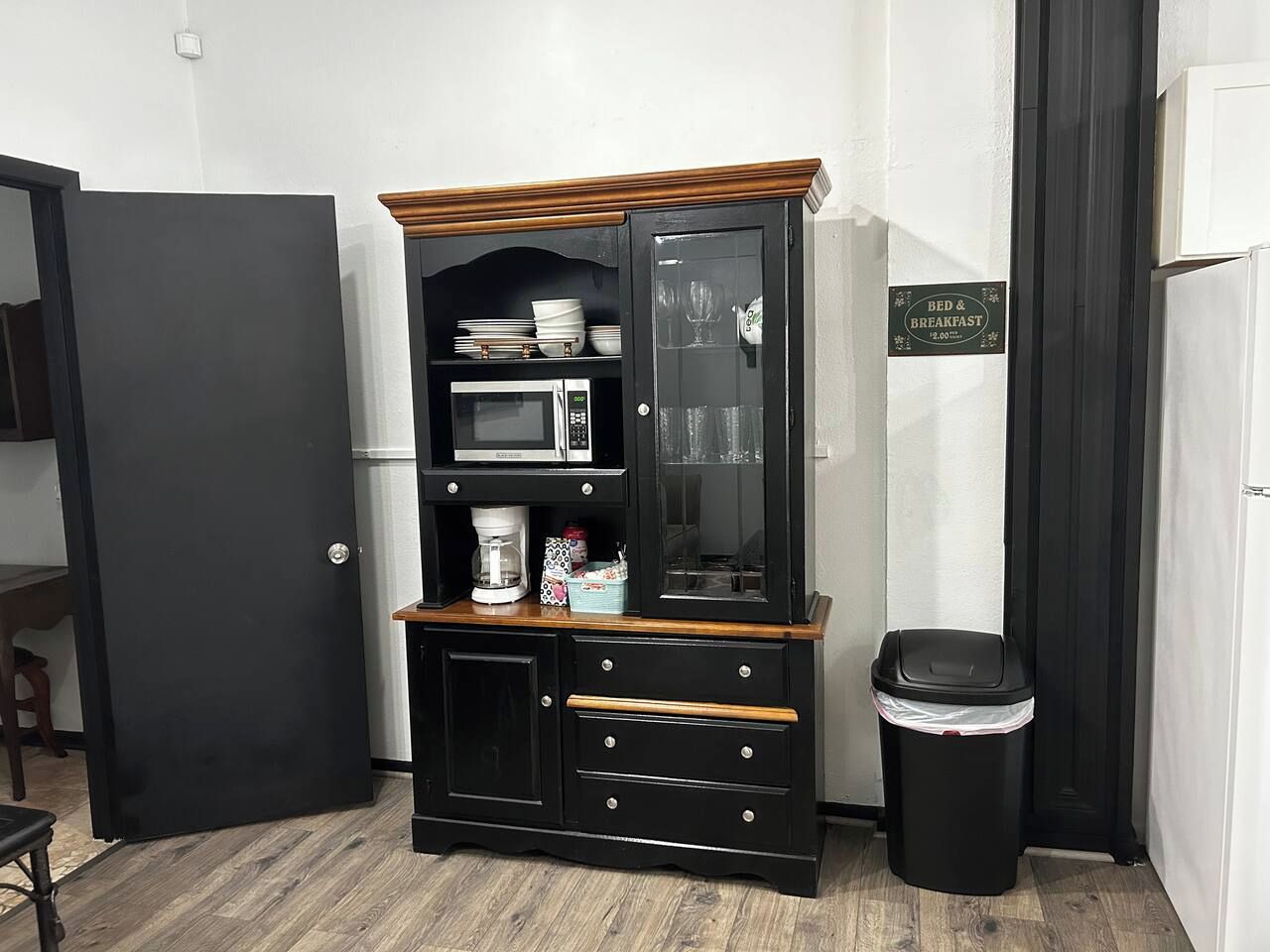Une petite cuisine équipée d’un four à micro-ondes, d’une machine à café et de tiroirs de rangement. On y trouve également une poubelle et un panneau indiquant « chambre d’hôtes ».