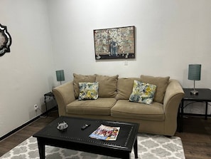 A living room with a beige sofa, two floral throw pillows, a coffee table with a magazine and remote control, a painting on the wall, and two lamps on side tables. - Kemp House Magnolia Suite | Historic Charm Downtown Ocala (Ocala)