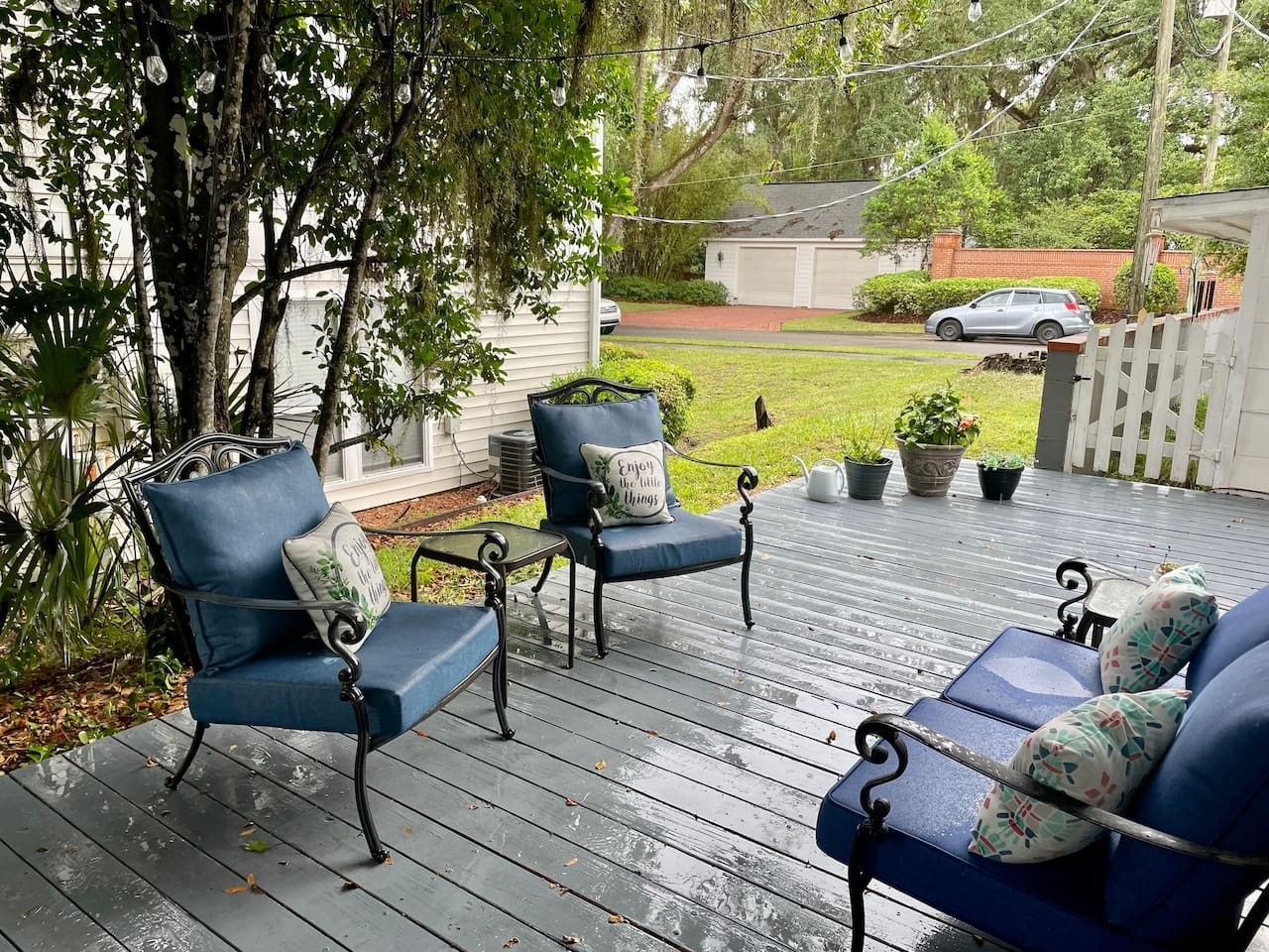 Une terrasse en bois, agrémentée de mobilier d’extérieur, notamment des chaises et un canapé, entourée de verdure et d’une clôture blanche.
