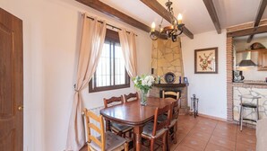 A dining area with a wooden table and chairs, a vase with flowers, a stone fireplace, and a kitchen area with a brick wall and a hanging light fixture.