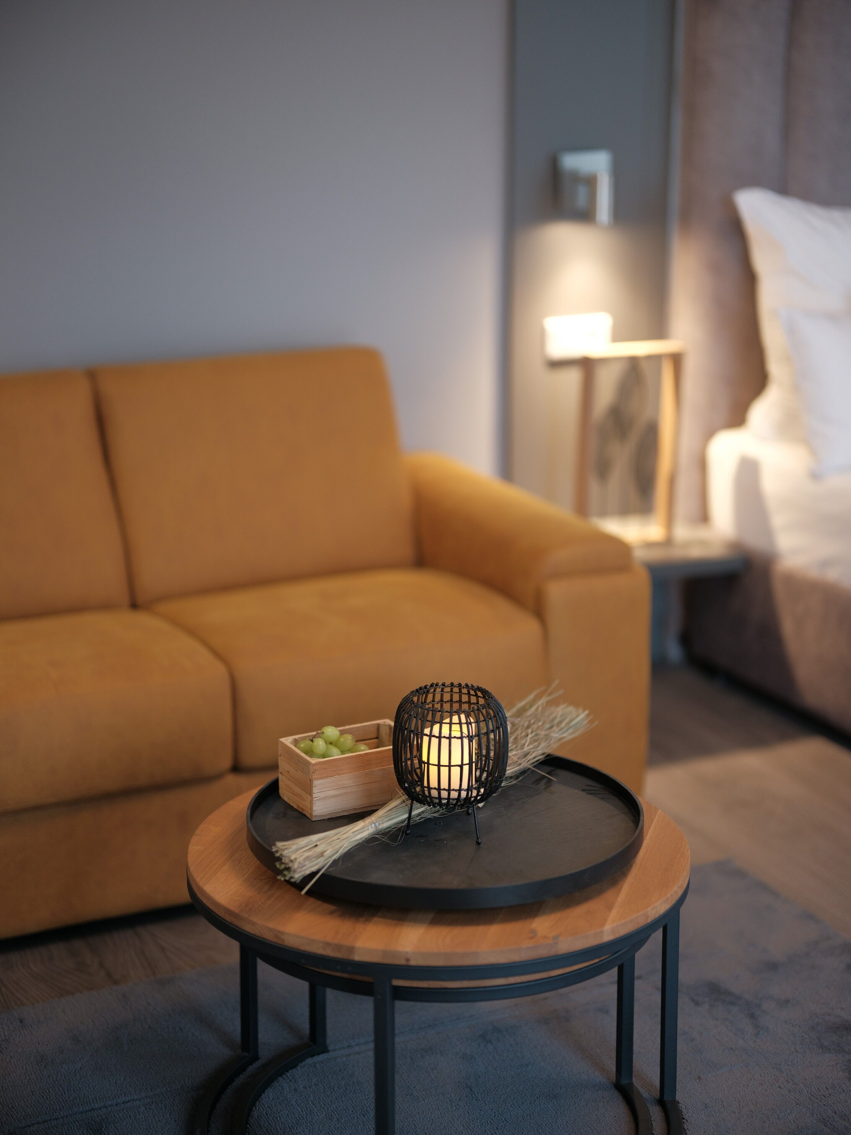 Una sala de estar moderna con un sofá, una mesa redonda de madera con un objeto de estructura metálica negra y un pequeño cofre de madera con uvas verdes.