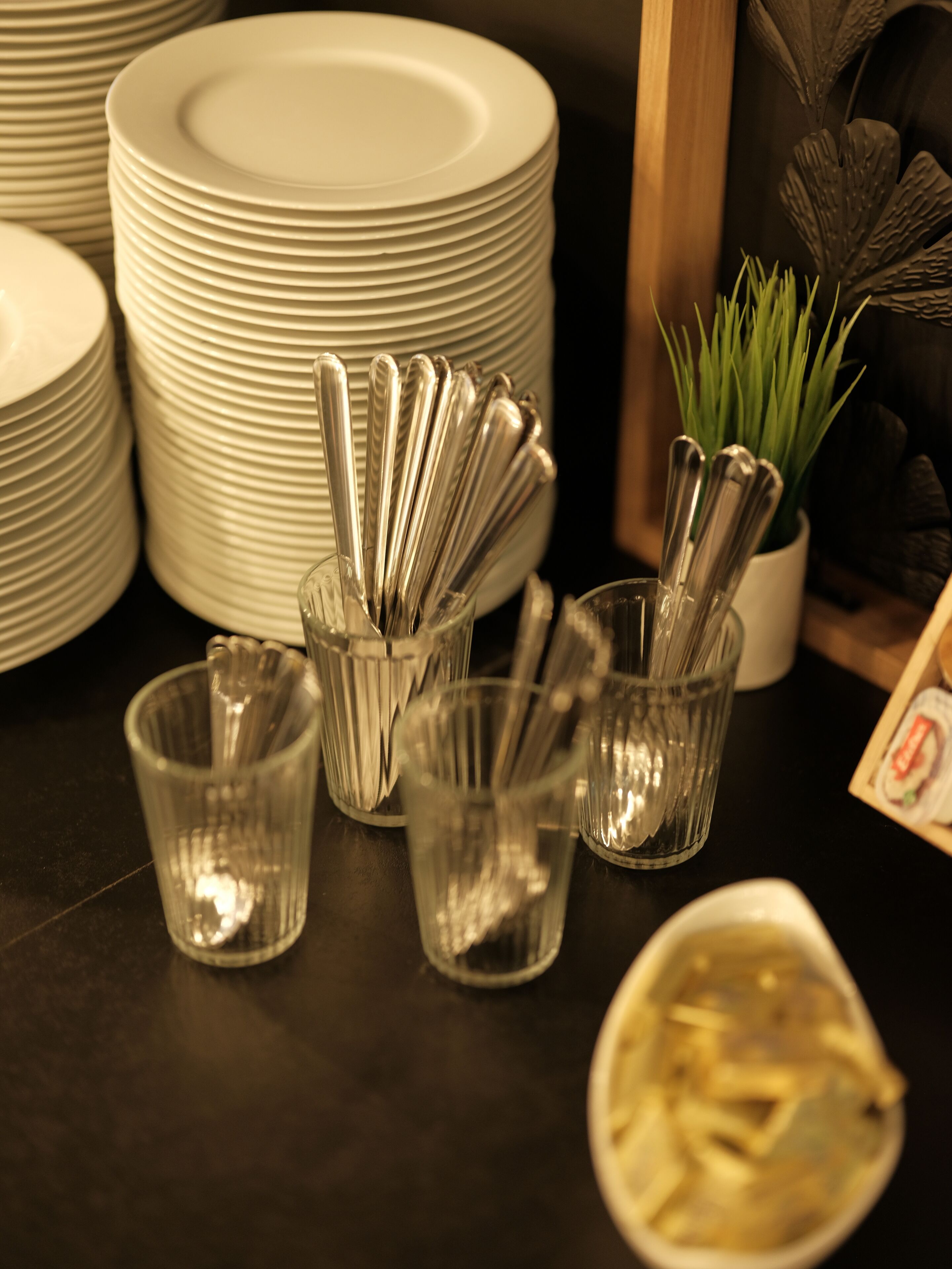 Ein Stapel weißer Teller, ein Besteckset in einem Glasbehälter und ein kleiner Blumentopf auf einer Tischplatte.