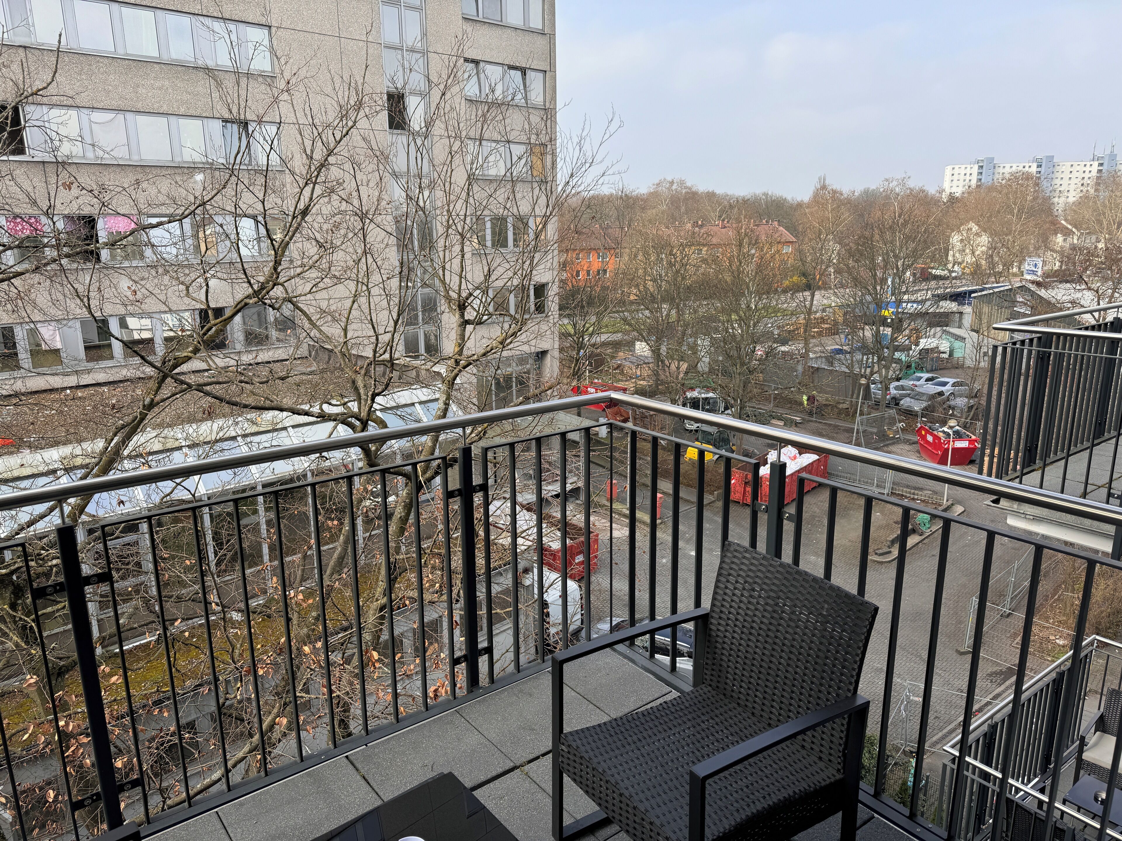 A balcony with a metal railing, a black woven chair, and a view of a parking area with cars and a red dumpster.