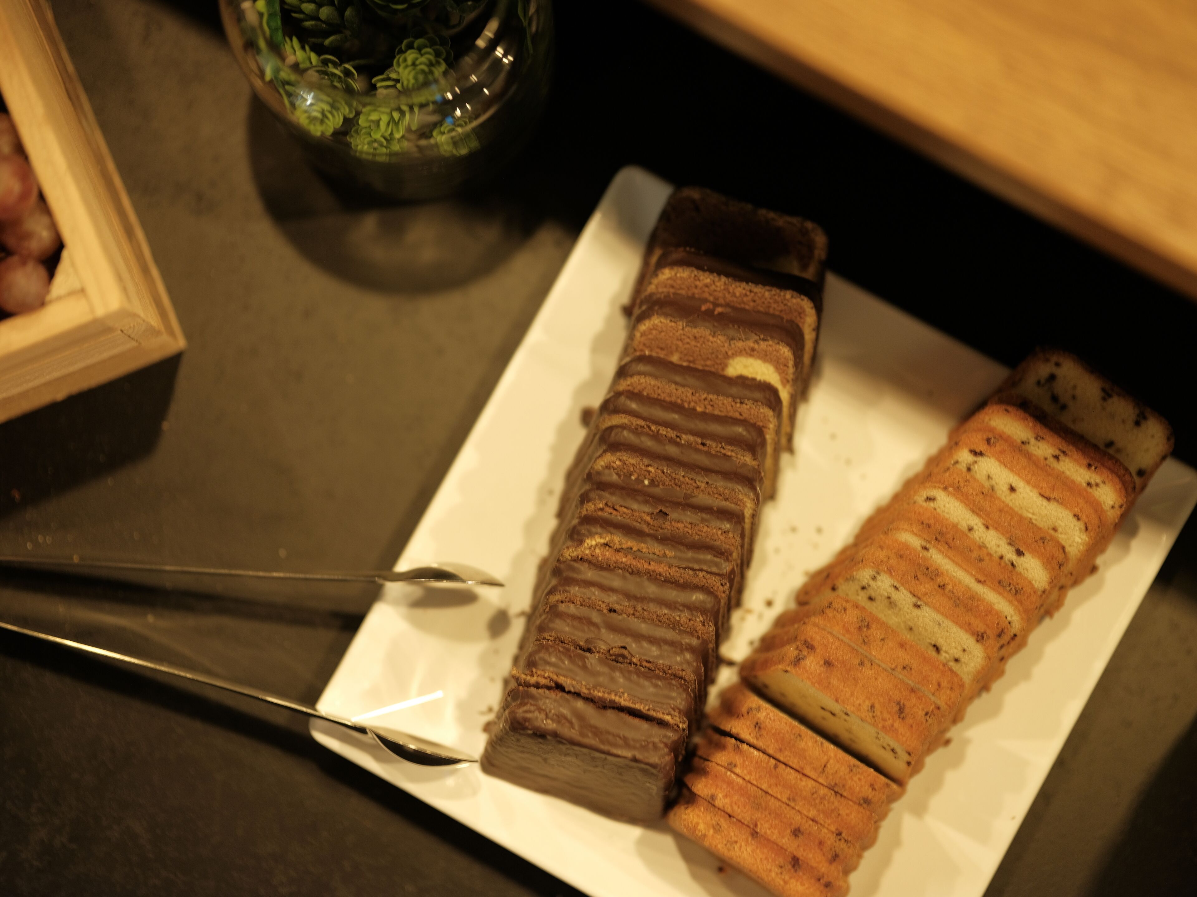 Ein weißer Teller mit zwei Kuchensorten, ein Schokoladenkuchen und ein Kuchen mit weißem Zuckerguss, auf einer dunklen Arbeitsplatte.