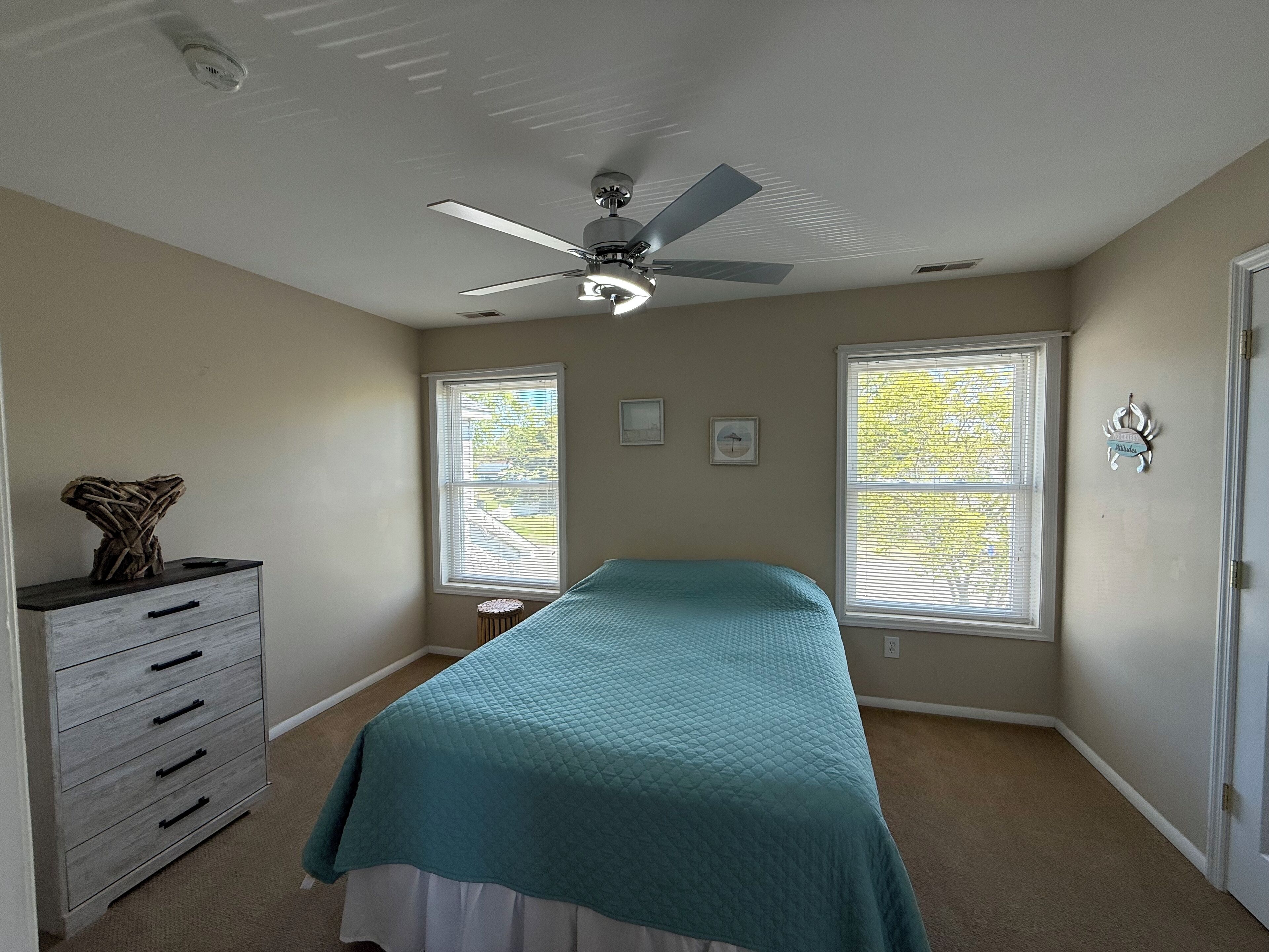 A bedroom with a bed, a ceiling fan, a dresser, and two windows with blinds.