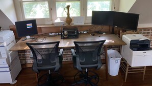 Um escritório em casa com duas mesas, duas cadeiras, dois monitores, uma impressora e um vaso no parapeito da janela.