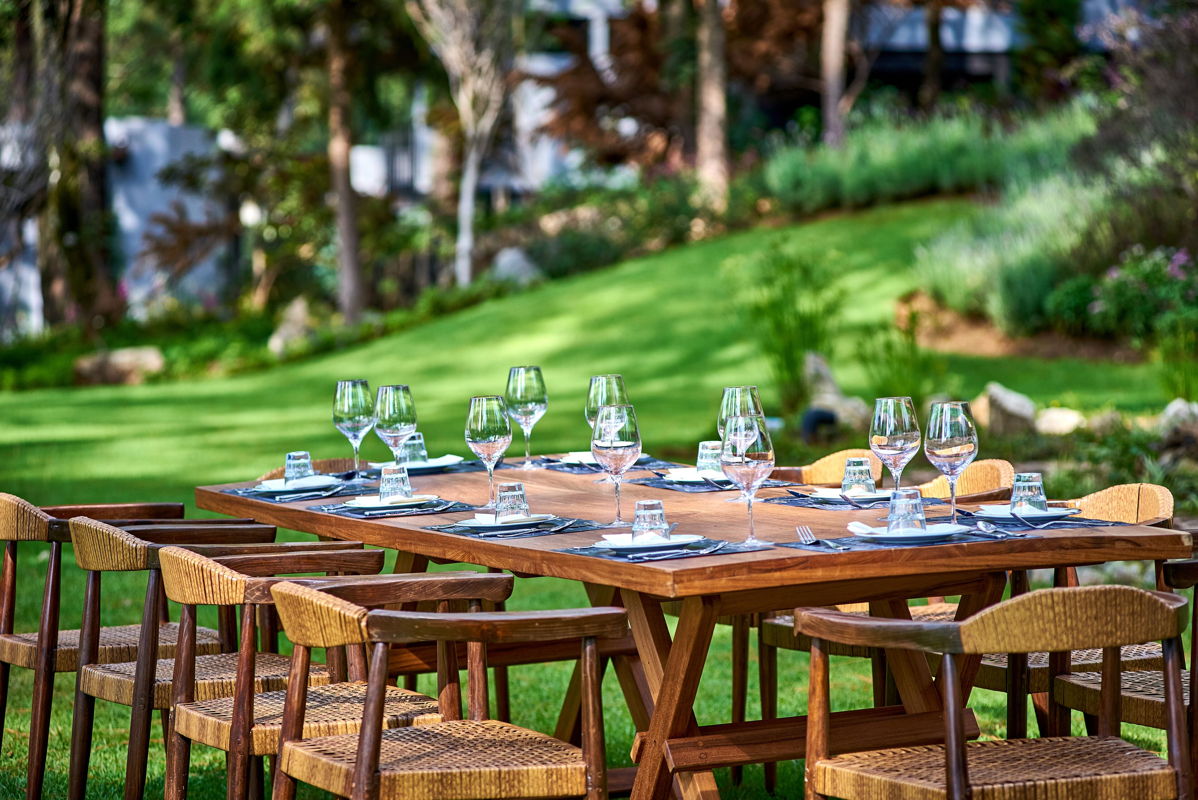 A wooden dining table set outdoors with wicker chairs, surrounded by greenery.