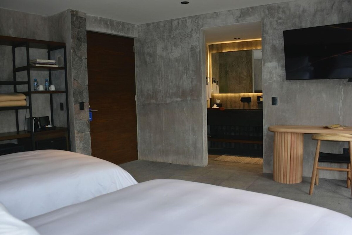 A modern hotel room with a large bed, a wooden side table, a TV mounted on the wall, and a bathroom visible through an open door.