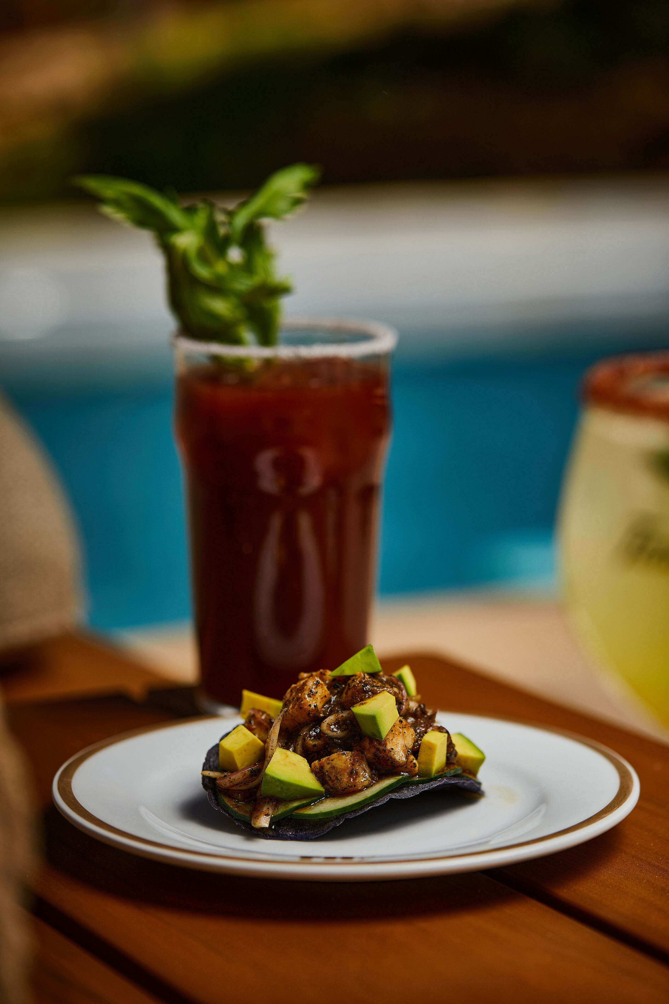 En tallerken med mad, avocado og kød, et glas rød drik og en pool i baggrunden.