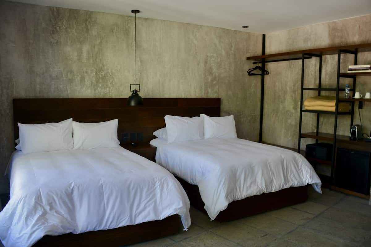 Ein Hotelzimmer mit zwei Einzelbetten, einem Holzkopfteil, einer Hängeleuchte und einem festen Regal mit einer Wasserflasche und einer Tasse.