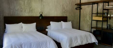 Une chambre d’hôtel avec deux lits simples, une tête de lit en bois, une lampe suspendue et une étagère intégrée où se trouvent une bouteille d’eau et une tasse.