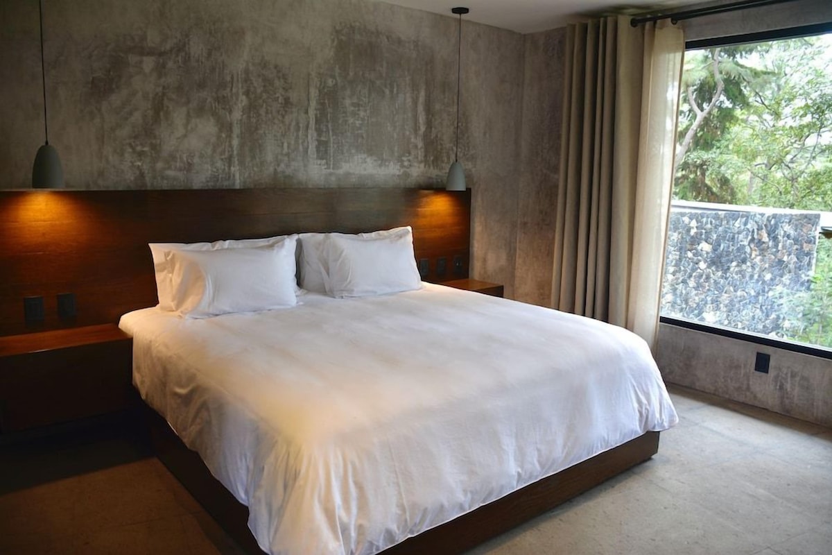 A large bed with a wooden headboard and two bedside lamps, a concrete wall, and a window with curtains.