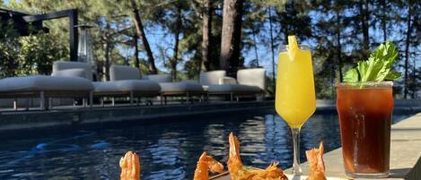 Una mesa de comedor con un plato de camarones, un vaso de jugo de naranja y un vaso de té helado junto a una piscina.