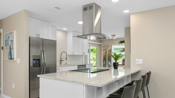 Een moderne keuken met witte kasten, een roestvrijstalen koelkast, een centraal keukenblok met barkrukken en een groot raam met uitzicht op groen.