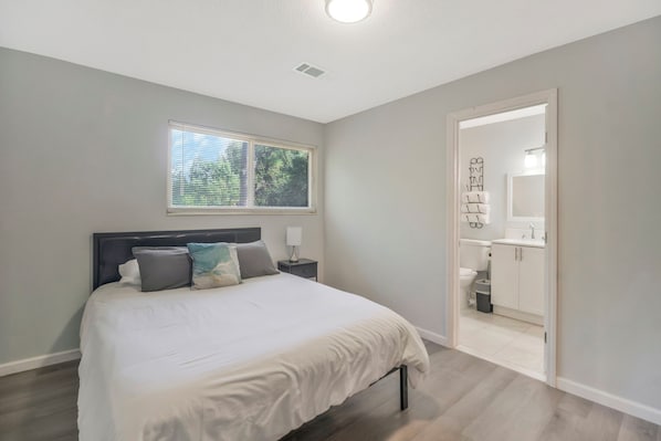 A bedroom with a bed, bedside tables, a window, and a bathroom visible through an open door.