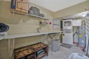 A laundry room with a washing machine, dryer, shelves with towels, and a trash can.