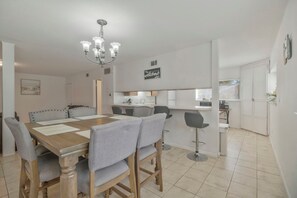 Une cuisine moderne avec des meubles blancs, un coin repas doté d’une table en bois et de chaises à revêtement gris, ainsi qu’un lustre.