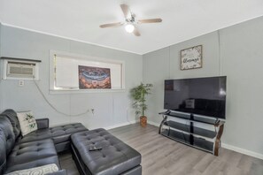 Un salon comprenant un canapé d’angle, un téléviseur à écran plat, un ventilateur de plafond, une fenêtre avec des stores et un cadre avec une citation accroché au mur.