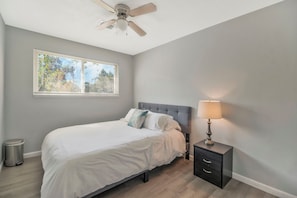 Une chambre à coucher comprenant un lit, une table de chevet, une lampe, un ventilateur de plafond et une fenêtre avec des stores.