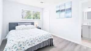 A bedroom with a bed, a nightstand, a window, and a bathroom with a sink and cabinets.