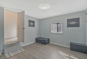 A room with a grey wall, a window with blinds, a grey ottoman, and a door leading to another room.