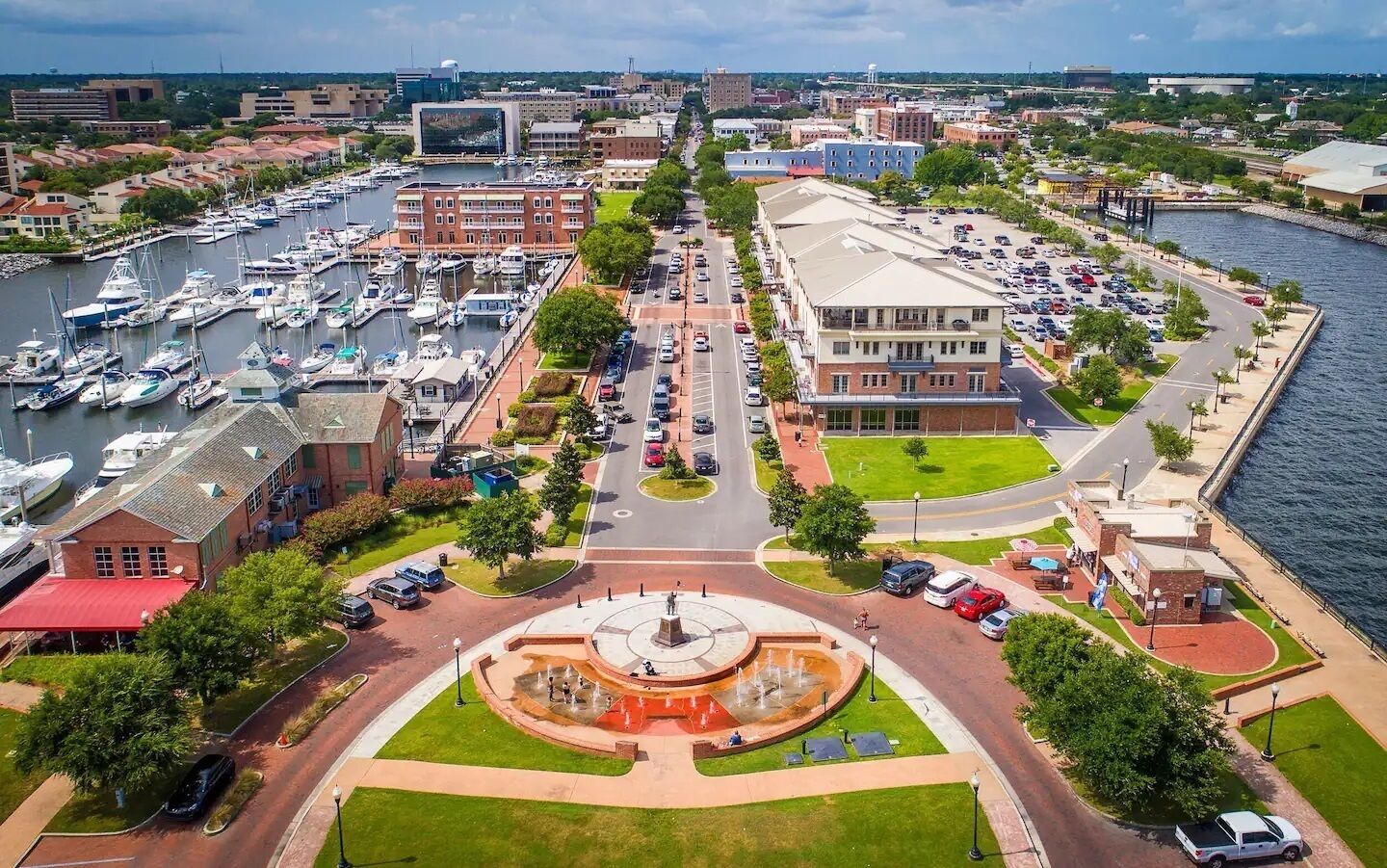 A marina with boats, a central fountain, and a building with a parking lot.