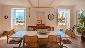 A dining area with a large table, four wicker chairs, a hanging light fixture, and a view of a cityscape through large windows. - Le Saint-Jean (Bastia)